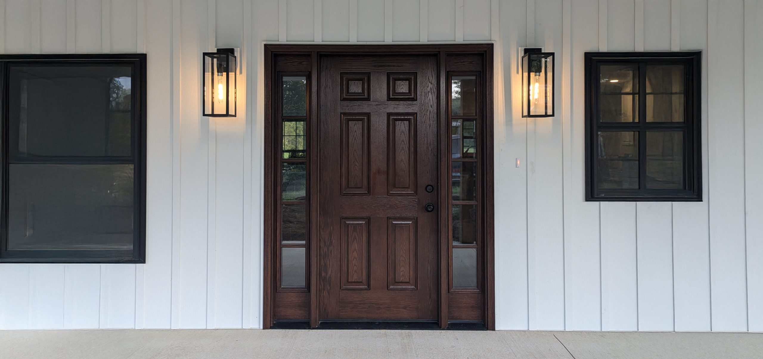 Front entrance of a home featuring a wooden door with decorative panels, flanked by sidelights and two black lantern-style wall sconces, set against a white exterior wall.